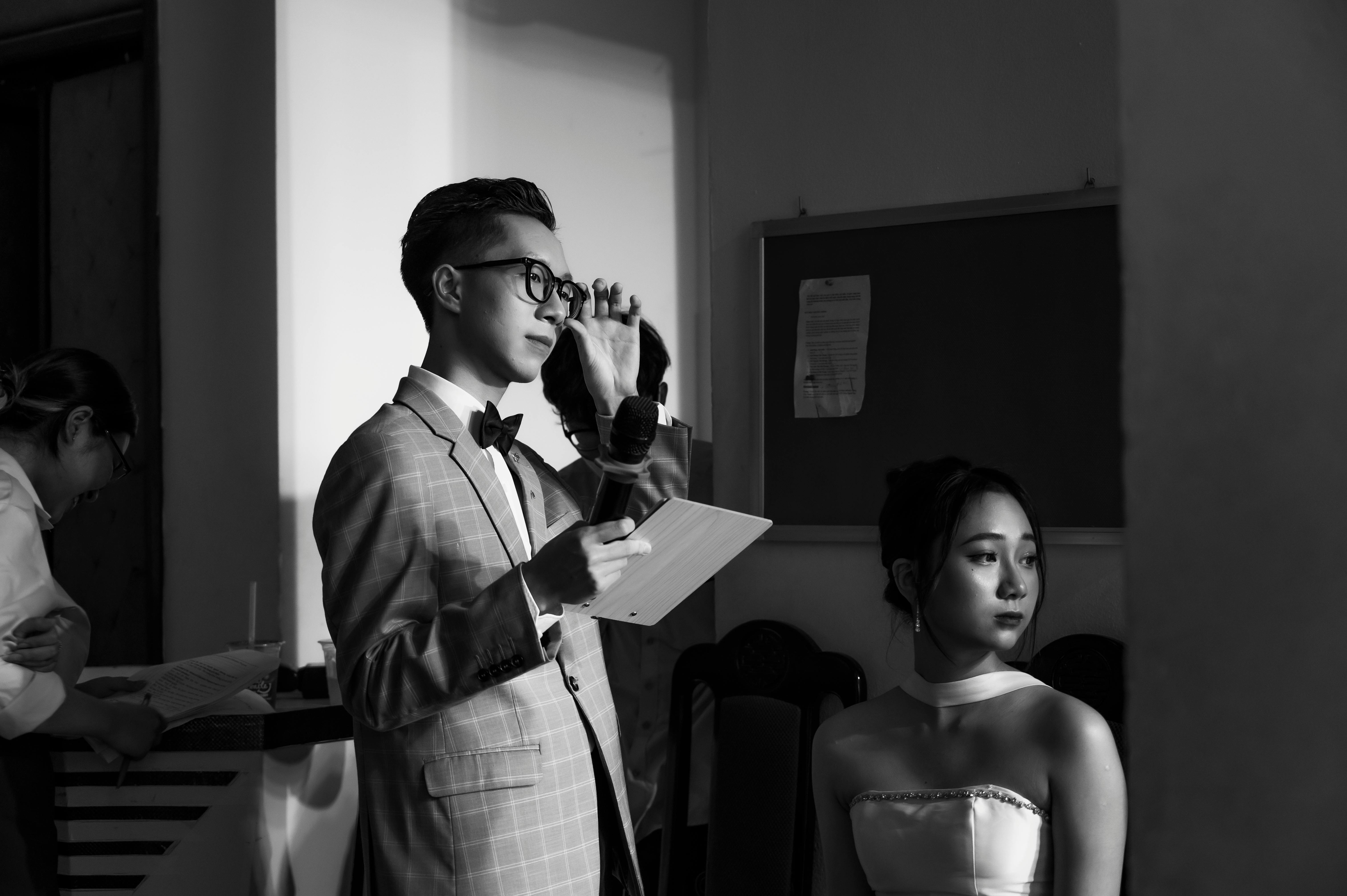 A man speaks into a microphone at an indoor event, elegantly dressed in a suit.